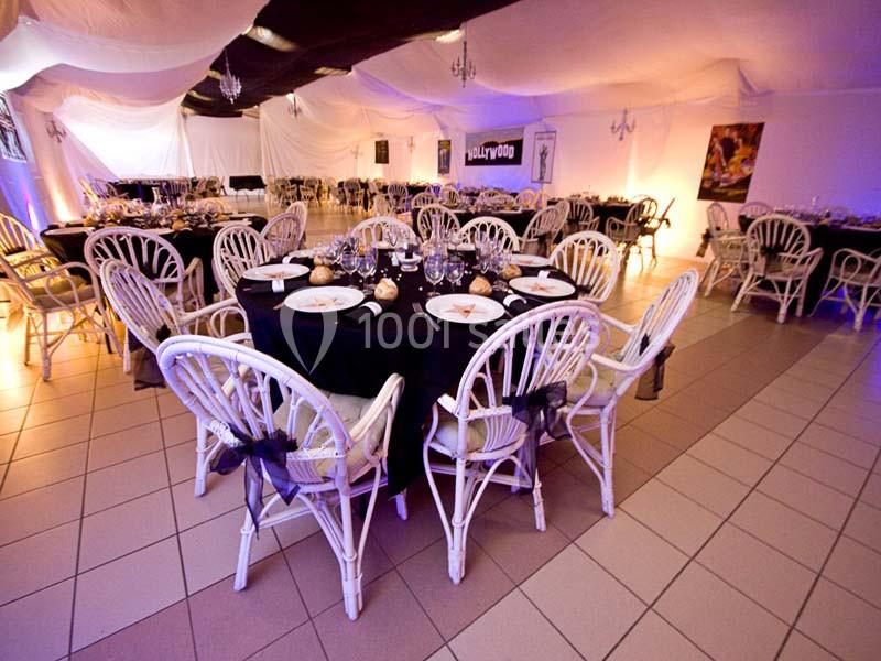 Salle de réception décorée avec des tables rondes dressées, chaises en osier blanc et éclairage tamisé.