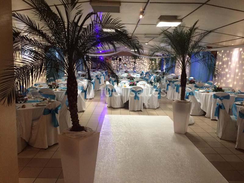 Salle de réception décorée avec des tables rondes, nappes blanches, nœuds bleus et palmiers en pots.