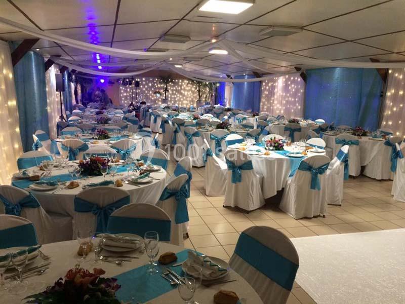 Salle de réception décorée avec des tables rondes, nappes blanches, nœuds bleus sur les chaises et éclairage tamisé.