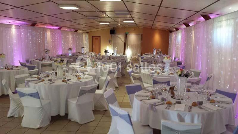 Salle de réception décorée avec des tables rondes, nappes blanches, chaises habillées et éclairage tamisé violet.