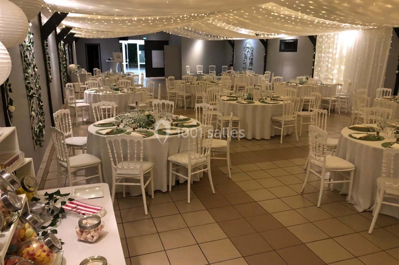Salle de réception décorée avec des tables rondes, nappes blanches, chaises élégantes et guirlandes lumineuses suspendues.