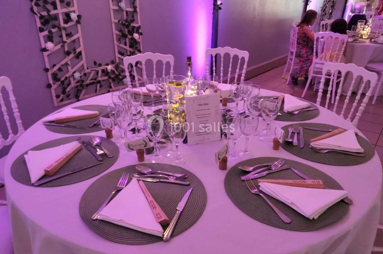 Table ronde dressée avec nappes blanches, couverts, verres, serviettes pliées et décoration lumineuse au centre.