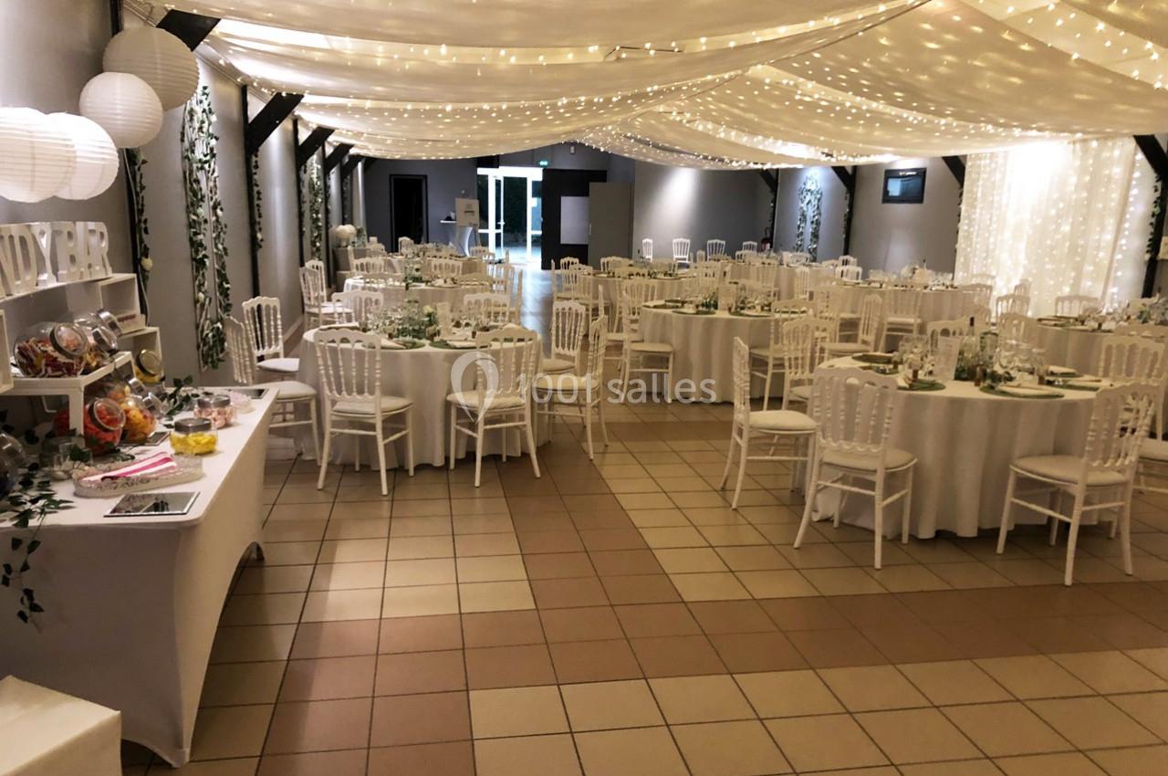 Salle de réception décorée avec des tables rondes, chaises blanches, guirlandes lumineuses et un buffet sur la gauche.