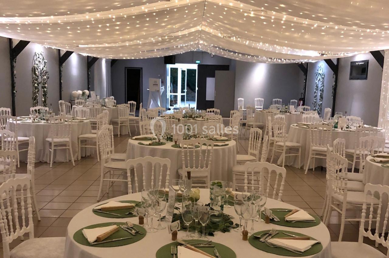 Salle de réception décorée avec des tables rondes, nappes blanches, chemins de table verts et guirlandes lumineuses…