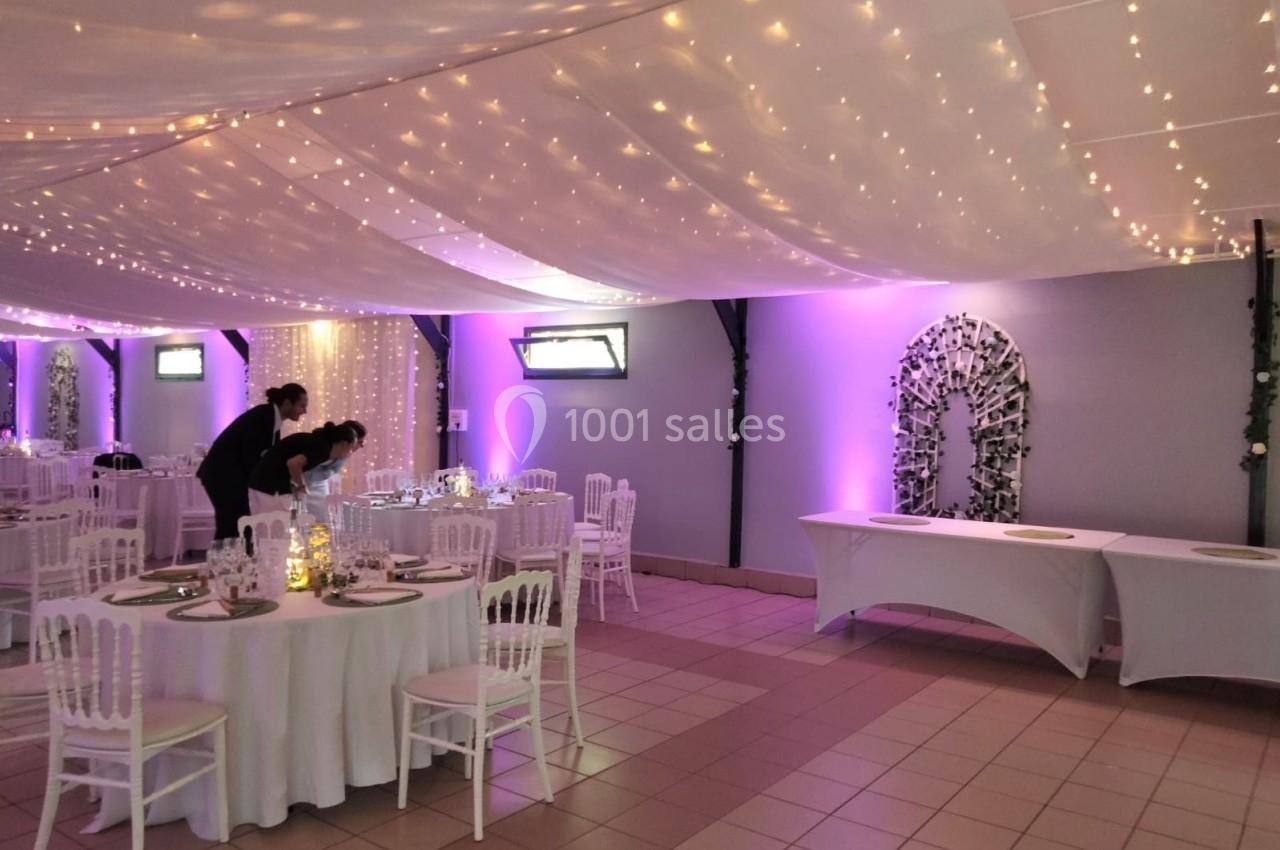 Salle de réception décorée avec des guirlandes lumineuses, tables rondes dressées et éclairage violet.