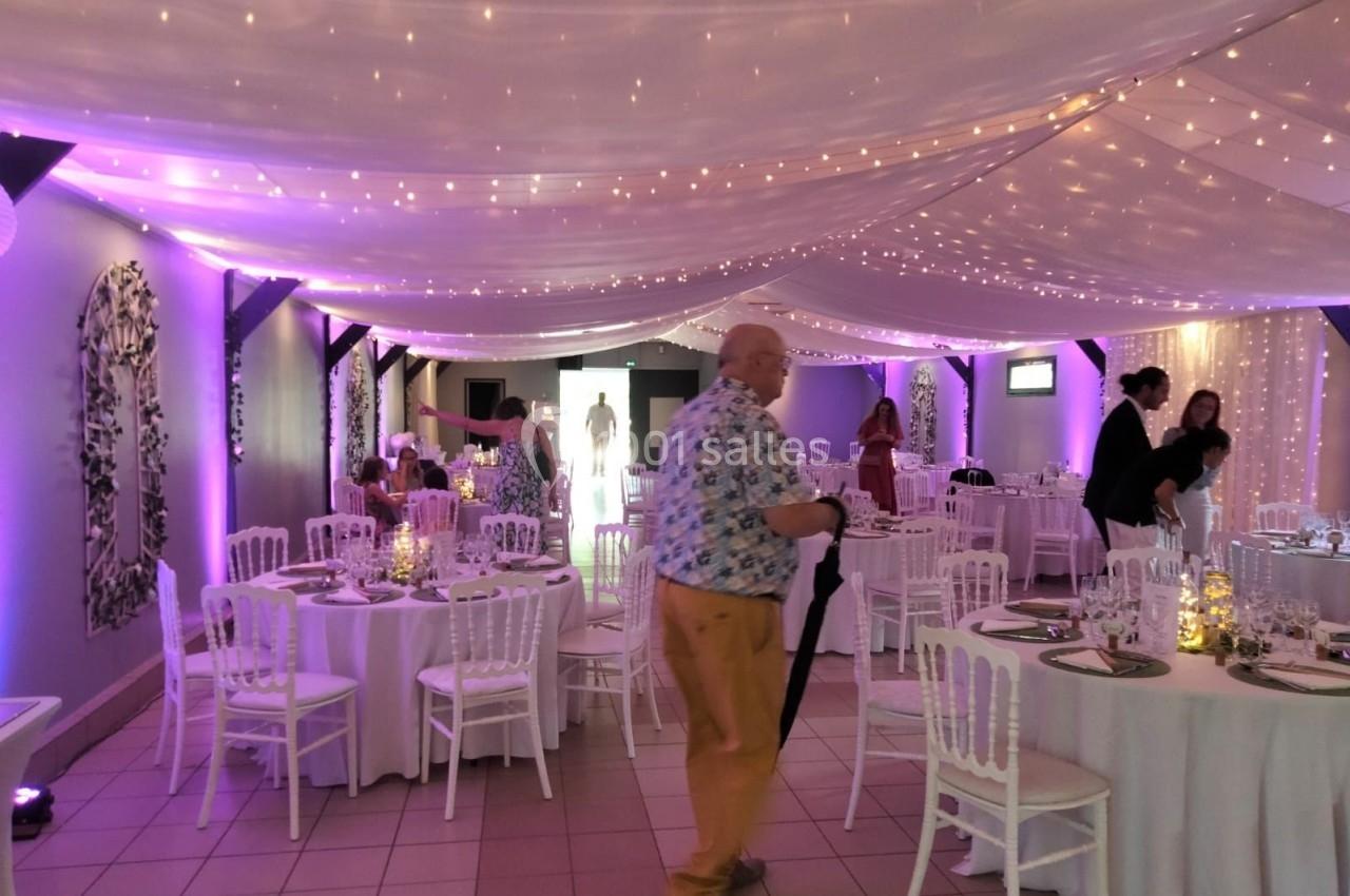 Salle de réception décorée avec des guirlandes lumineuses, tables rondes dressées et chaises blanches.