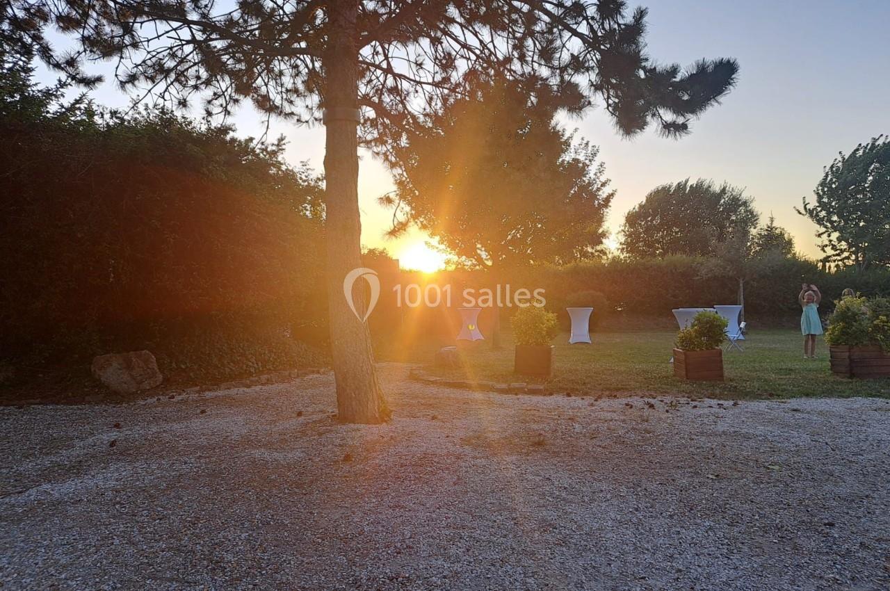 Coucher de soleil illuminant un jardin avec un arbre au premier plan et des tables hautes en arrière-plan.