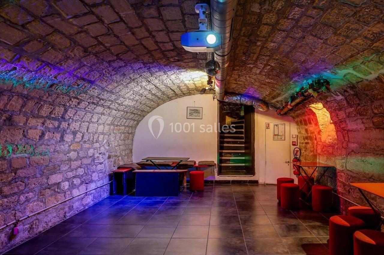 Salle voûtée en pierre avec éclairage coloré, mobilier moderne et escalier menant à une porte au fond.