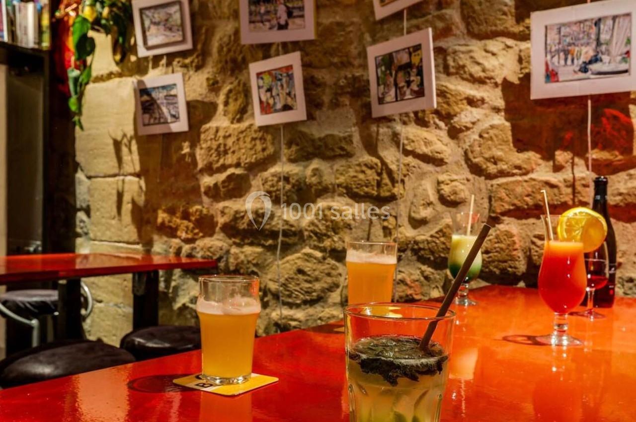 Tables rouges avec cocktails variés dans un bar à l'ambiance chaleureuse, murs en pierre décorés de photos.