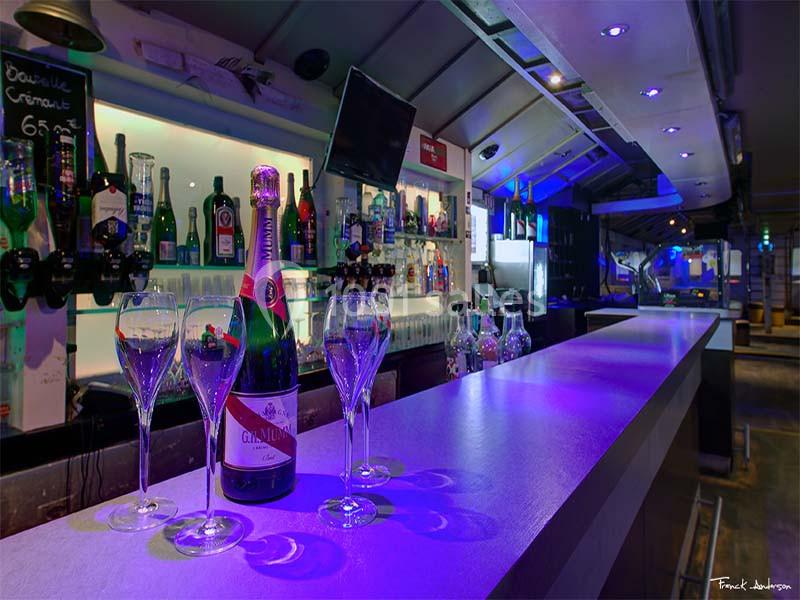 Bar moderne avec comptoir éclairé, bouteilles en arrière-plan, trois verres et une bouteille de champagne au premier plan.