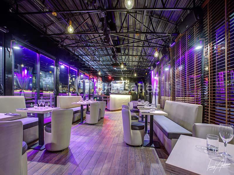 Salle de restaurant moderne avec tables dressées, banquettes blanches, éclairage tamisé et grandes baies vitrées.