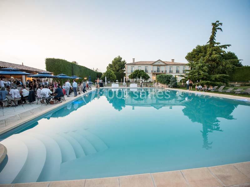 Piscine extérieure entourée de chaises longues, avec une grande maison et des invités en arrière-plan.