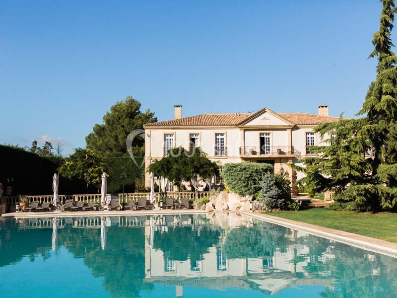 Grande maison en pierre avec piscine au premier plan, entourée de végétation et sous un ciel bleu clair.