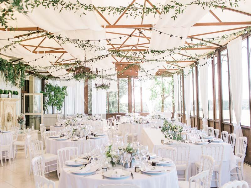 Salle de réception lumineuse décorée de guirlandes végétales, tables rondes dressées et chaises blanches élégantes.