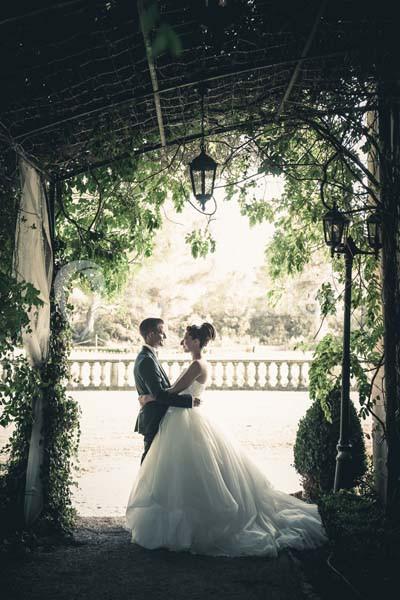 Un couple en tenue de mariage se tient sous une arche végétale, baigné par une lumière douce.