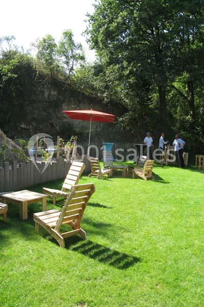 Chaises en bois et tables disposées sur une pelouse avec un parasol rouge, entourées d'arbres et d'une clôture en bois.