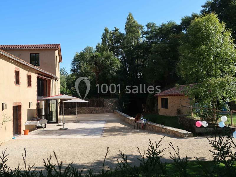 Cour extérieure ensoleillée avec terrasse, parasol, maison en pierre, arbres en arrière-plan et quelques ballons colorés.