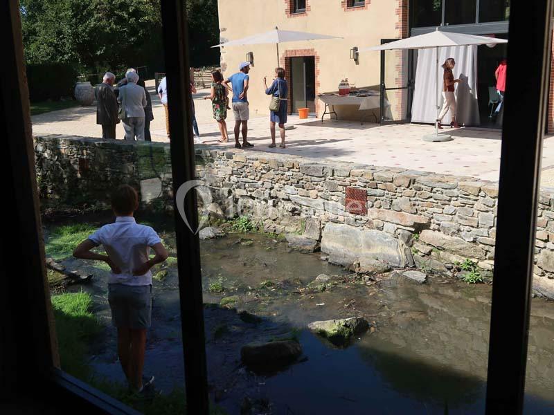 Un enfant observe un ruisseau depuis une fenêtre, tandis que des adultes discutent près d'un bâtiment en arrière-plan.
