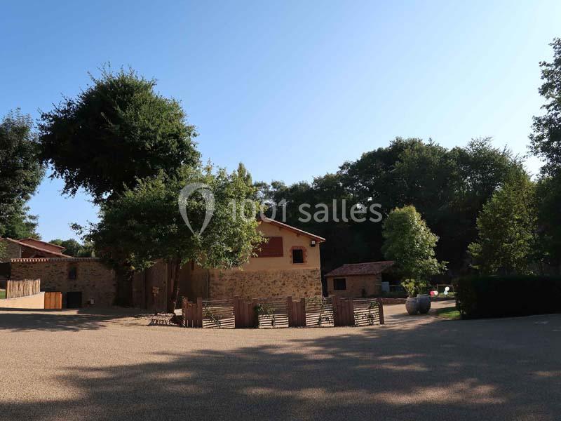 Cour ensoleillée avec une maison en pierre, des arbres, une clôture en bois et un chemin en gravier.