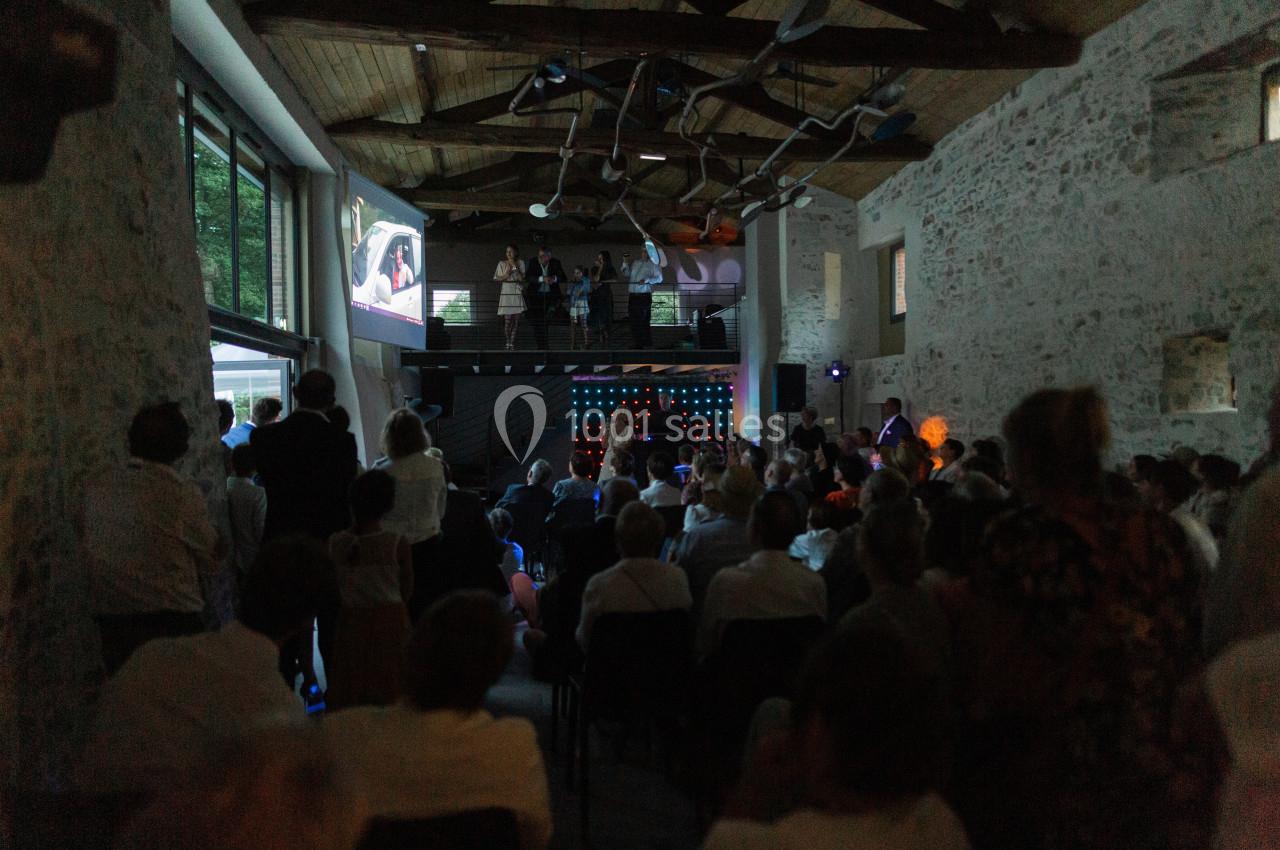 Salle en pierre avec des invités assis, regardant une projection et une scène éclairée au fond.