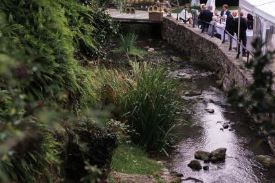 Petit ruisseau bordé de végétation avec des tentes blanches installées pour un événement en arrière-plan.