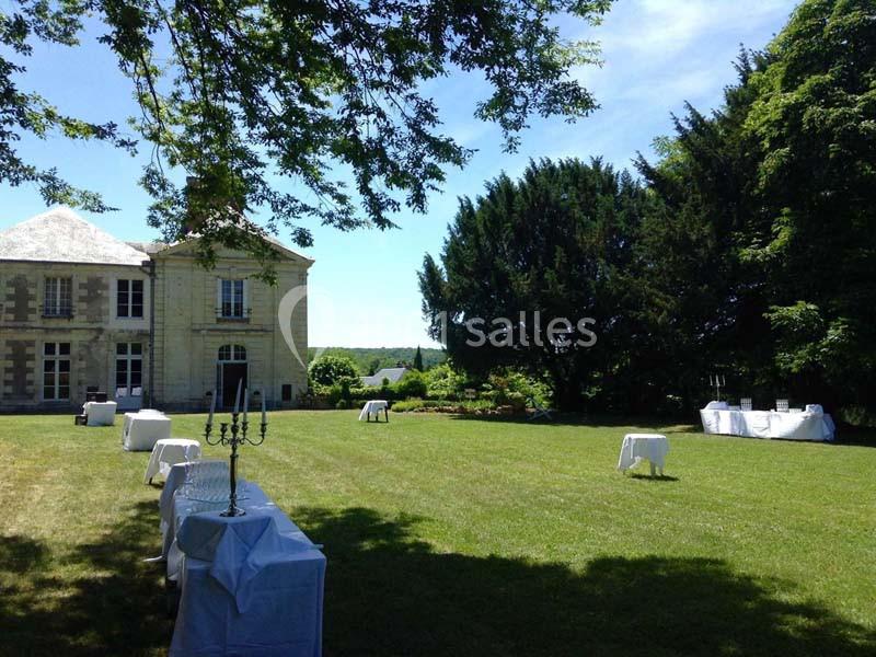 Jardin d'un domaine avec pelouse aménagée pour un événement, tables dressées et bâtiment en pierre à gauche.