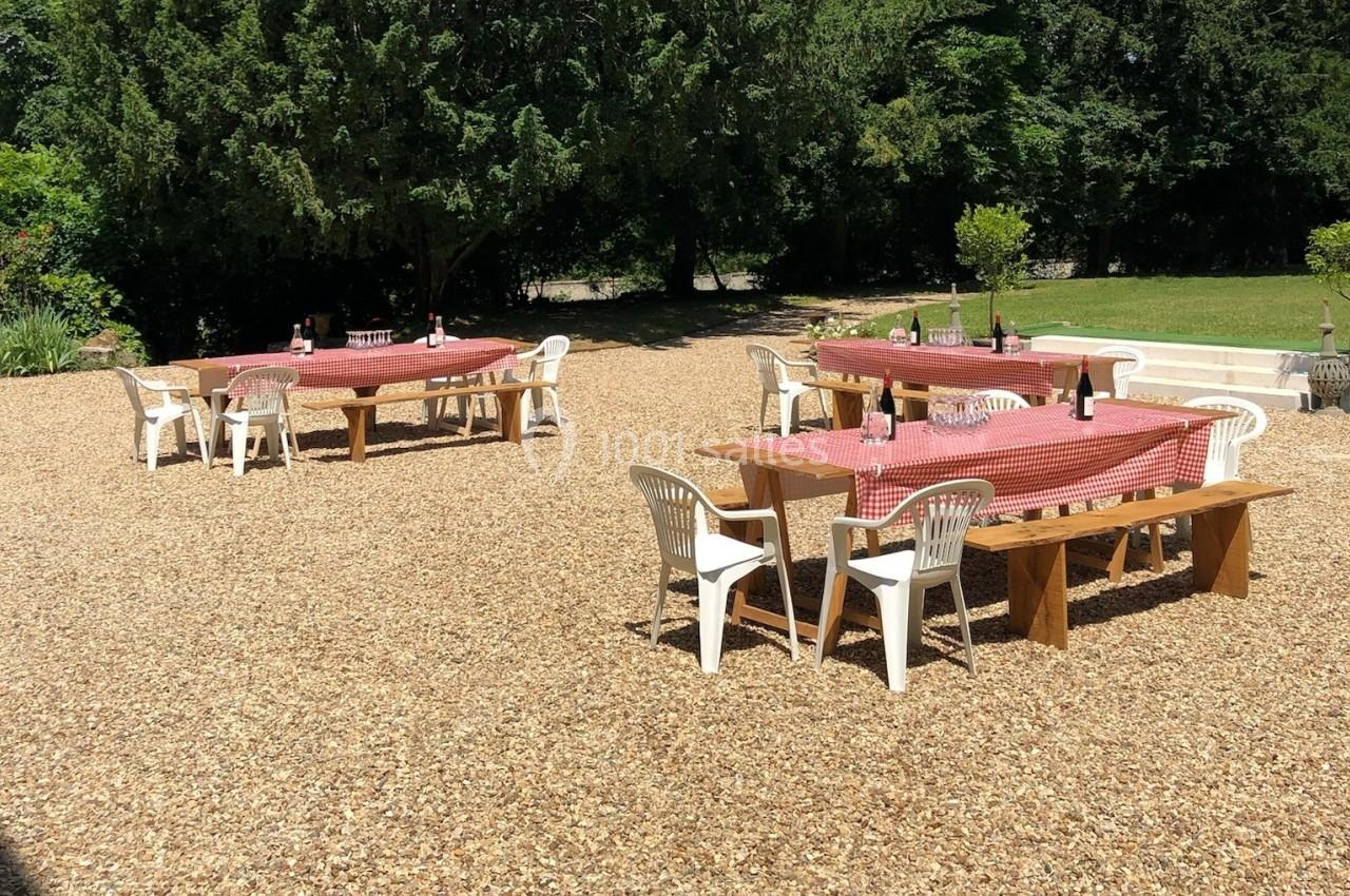 Tables en bois avec nappes rouges et chaises blanches disposées sur un sol gravillonné, entourées de verdure.