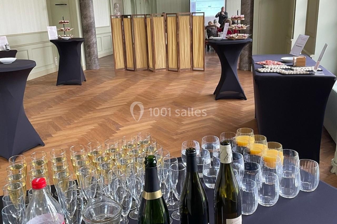 Salle avec parquet, tables hautes noires, boissons, verres et buffet disposés pour un événement ou une réception.
