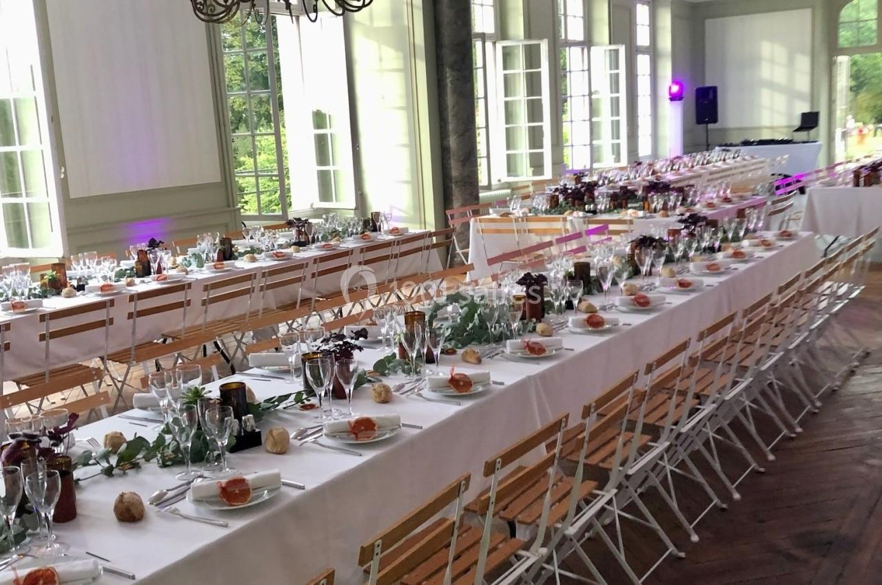 Salle de réception lumineuse avec de longues tables décorées de nappes blanches, vaisselle et éléments végétaux.