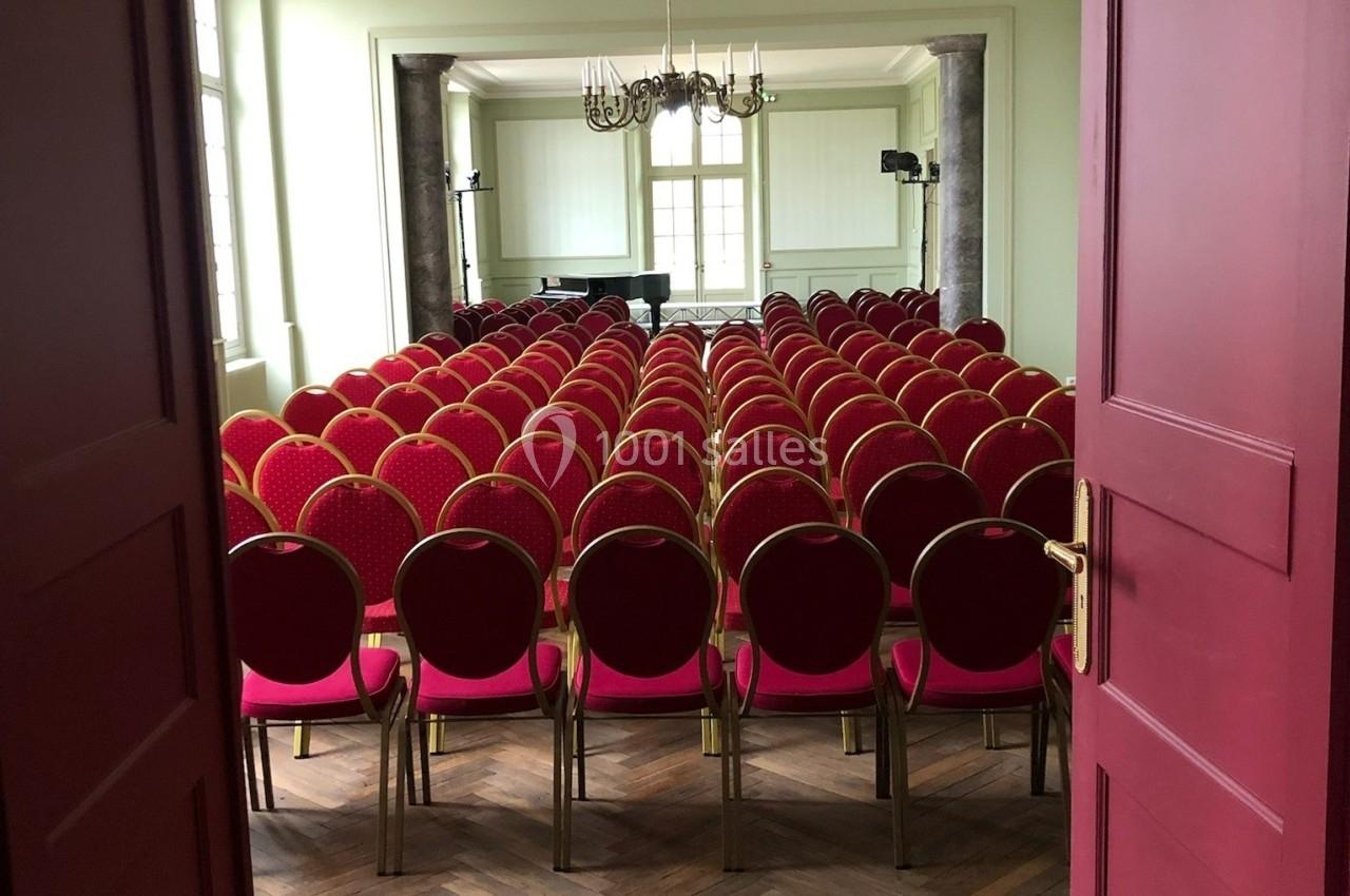 Salle de conférence avec des rangées de chaises rouges alignées face à un piano et un chandelier au plafond.