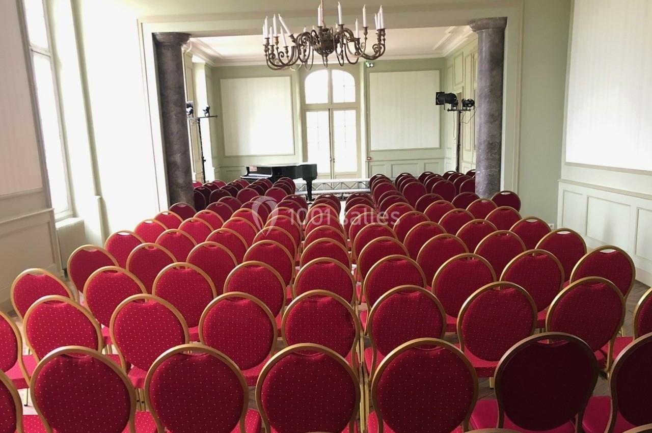 Salle de conférence avec des rangées de chaises rouges alignées face à un piano et une grande fenêtre.