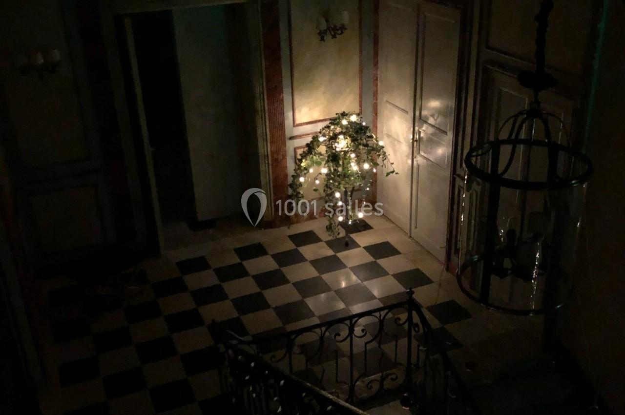 Hall d'entrée sombre avec un sol en damier, un escalier en fer forgé et un arbre décoré de lumières.