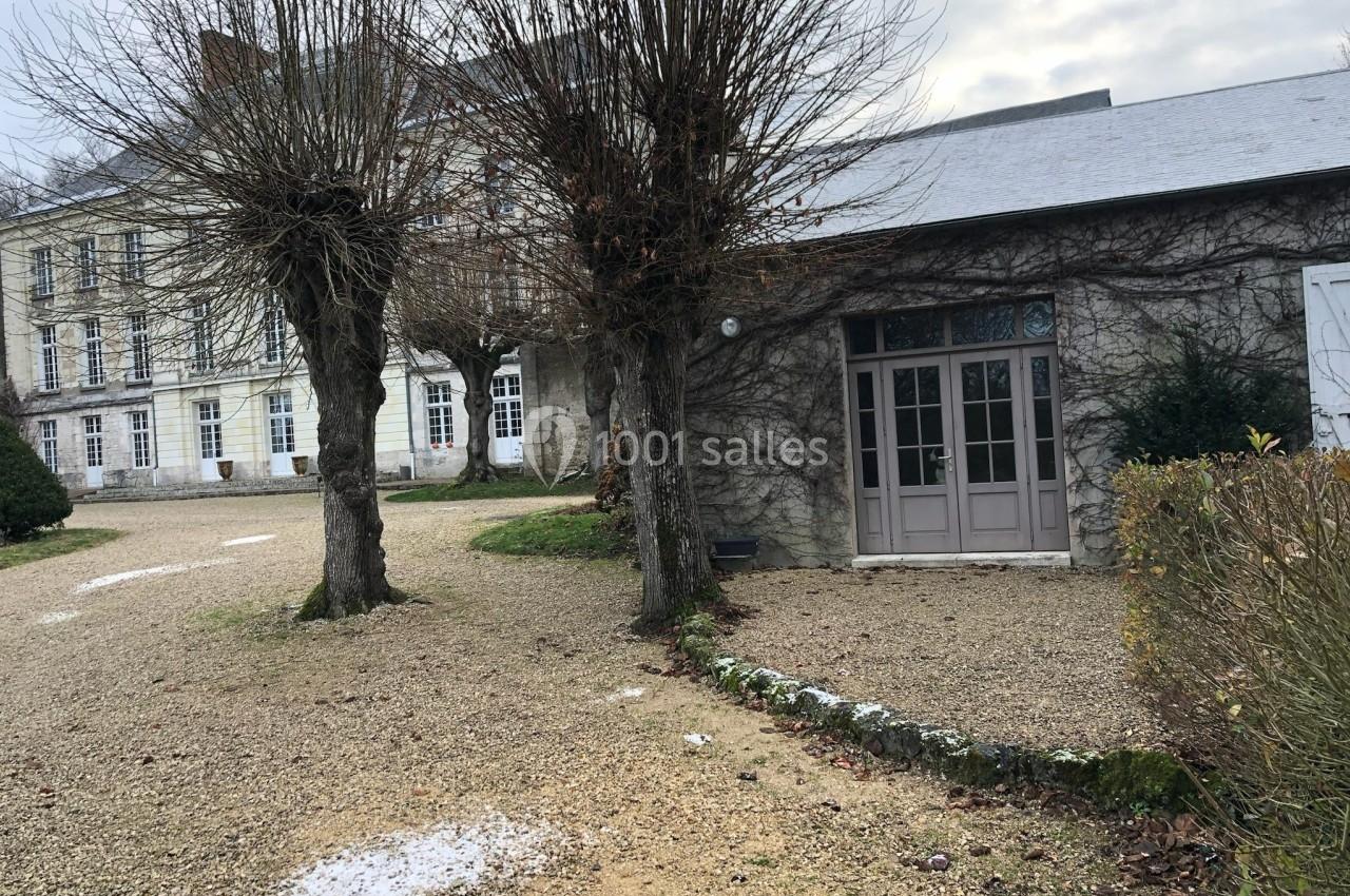 Bâtiment en pierre avec porte vitrée, arbres dénudés et gravier au sol, sous un ciel nuageux.