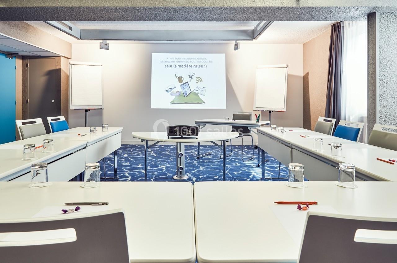 Salle de réunion lumineuse avec tables disposées en U, projecteur, écran, et tableaux blancs.
