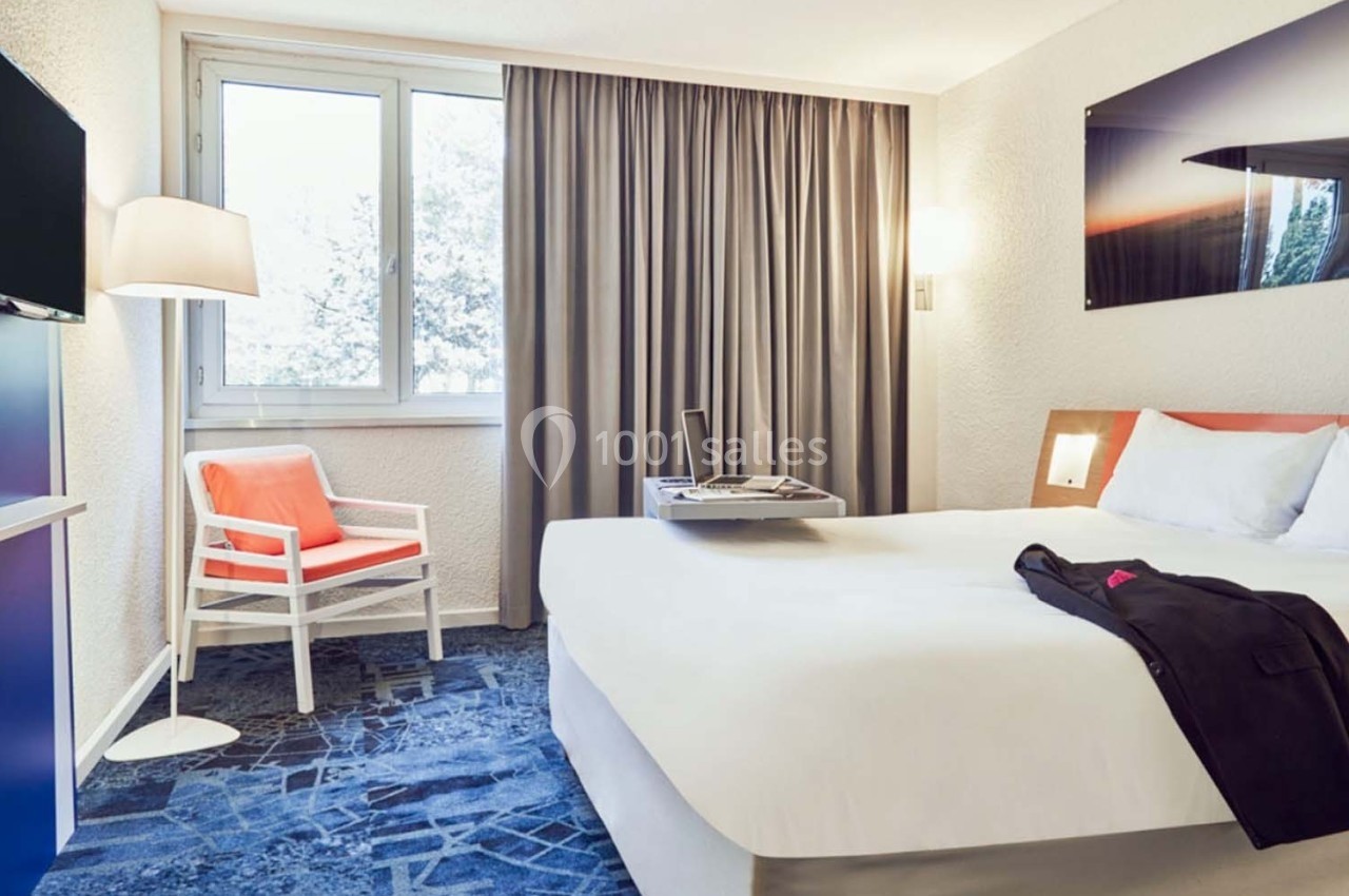 Chambre d'hôtel lumineuse avec un lit double, un fauteuil, une télévision, une fenêtre et des rideaux gris.