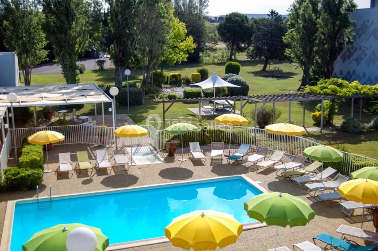 Piscine extérieure entourée de transats et parasols colorés, avec un jardin arboré en arrière-plan.