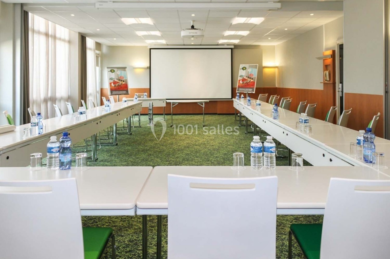 Salle de réunion lumineuse avec tables disposées en U, chaises blanches, écran de projection et bouteilles d'eau.