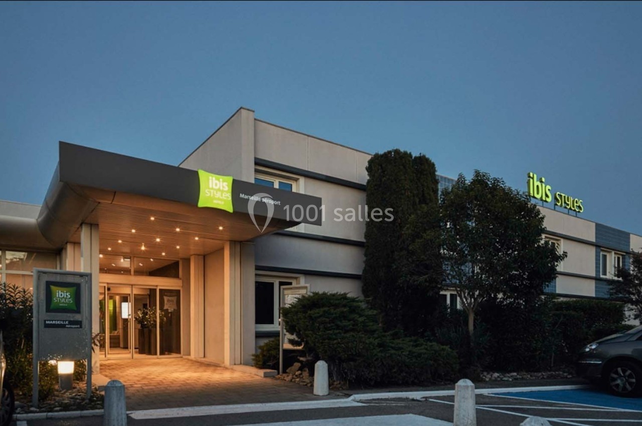 Façade d'un hôtel Ibis Styles en soirée, avec enseigne lumineuse et entrée éclairée entourée de végétation.