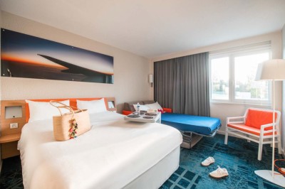 Chambre d'hôtel lumineuse avec lit double, canapé-lit, table basse, fenêtre et décoration moderne.