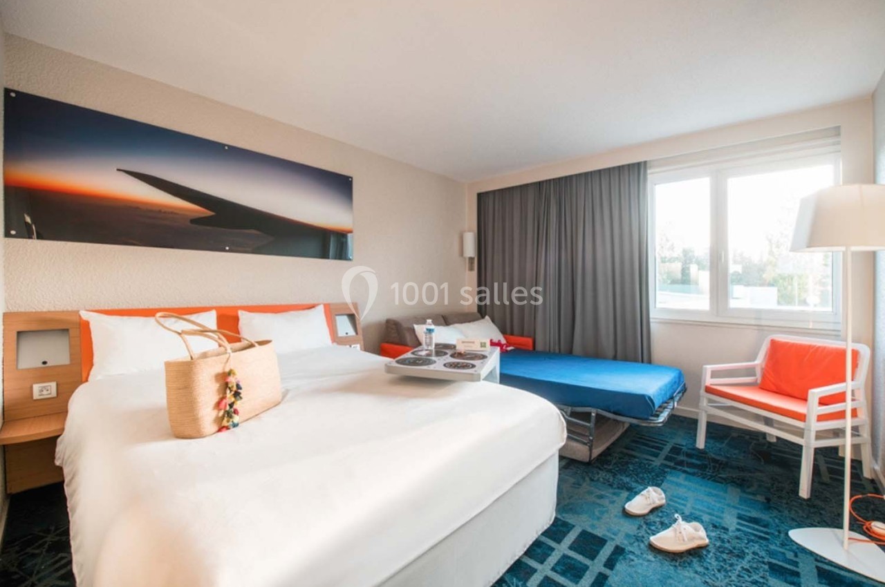 Chambre d'hôtel lumineuse avec lit double, canapé-lit, table basse, fenêtre et décoration moderne.