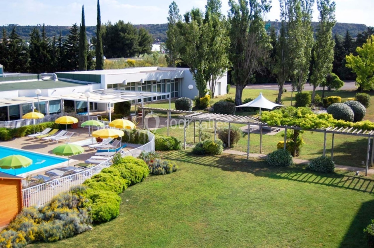 Vue d'un jardin avec piscine, chaises longues, parasols colorés et pergola entourés de végétation.