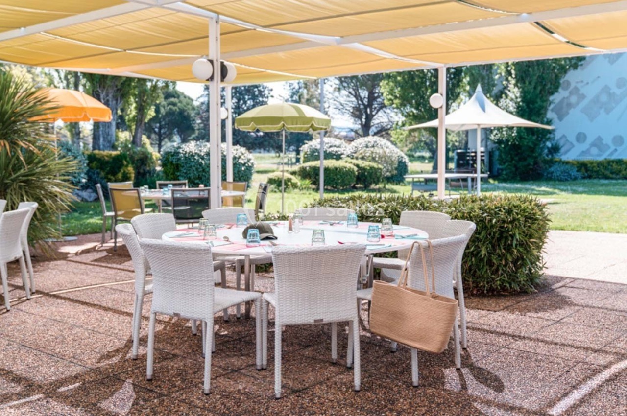 Table en plein air avec chaises blanches sous des parasols colorés, entourée de verdure et d'arbustes.