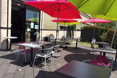 Terrasse en bois avec tables, chaises colorées et parasols verts et rouges, entourée de végétation et de bâtiments.