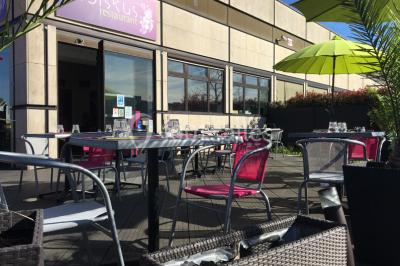 Terrasse en bois avec tables, chaises colorées et parasols verts et rouges, entourée de végétation et de bâtiments.