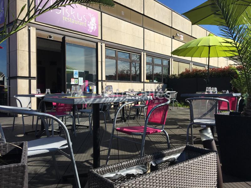 Terrasse d'un restaurant avec tables et chaises en métal, parasols verts et plantes sous un ciel ensoleillé.