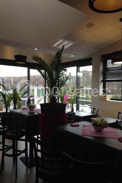 Salle de restaurant lumineuse avec des tables noires, des plantes décoratives et de grandes baies vitrées.