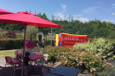 Terrasse en bois avec tables, chaises colorées et parasols verts et rouges, entourée de végétation et de bâtiments.