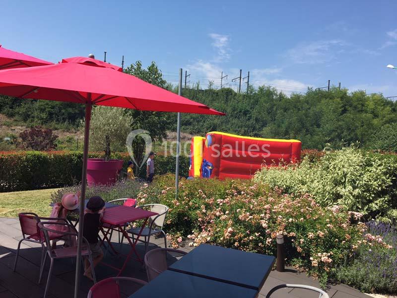 Terrasse avec tables et parasols rouges, vue sur un jardin fleuri et un château gonflable coloré en arrière-plan.