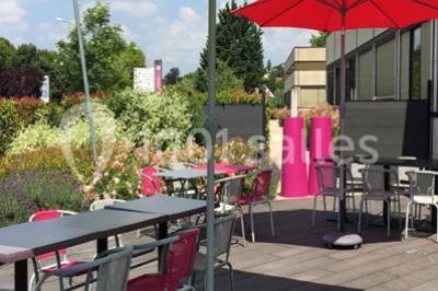 Terrasse en bois avec tables, chaises colorées et parasols verts et rouges, entourée de végétation et de bâtiments.