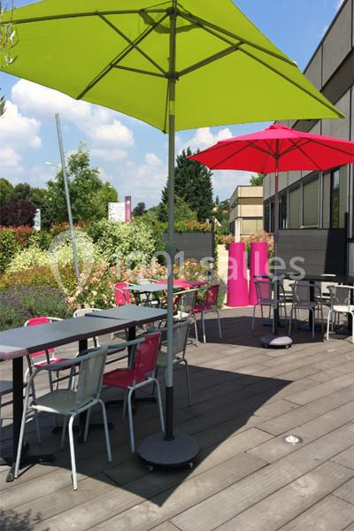 Terrasse en bois avec tables, chaises colorées et parasols verts et rouges, entourée de végétation et de bâtiments.